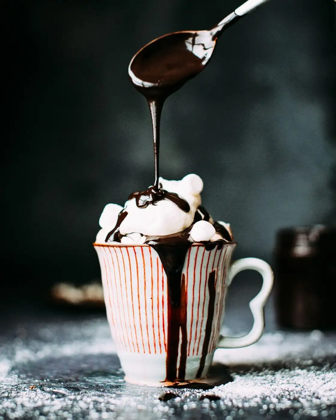 Thick chocolate sauce running off a spoon onto whipped cream and marshmallows piled over a striped mug