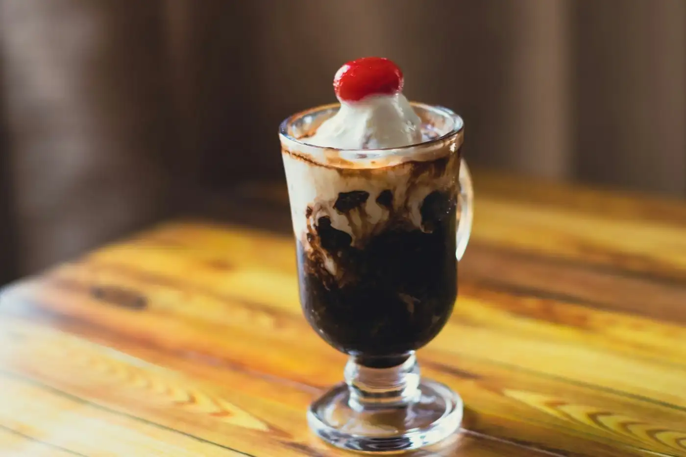 A hot fudge sundae in a footed glass mug, vanilla ice cream marbled with fudge and a red cherry on top, on a warm wooden counter