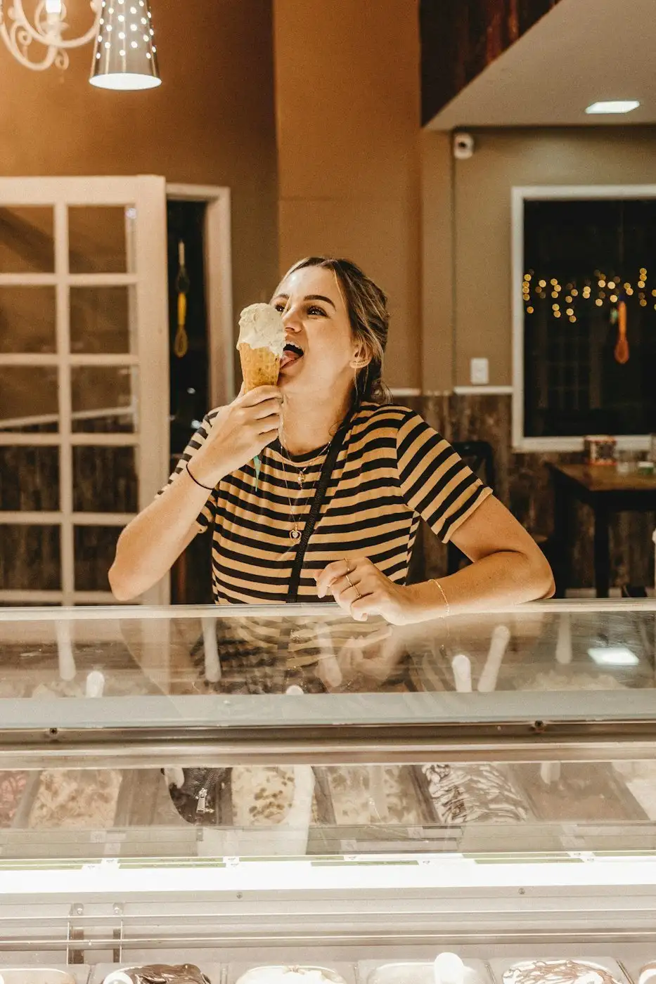 A smiling woman in a striped shirt tastes a waffle cone behind the glass dipping case