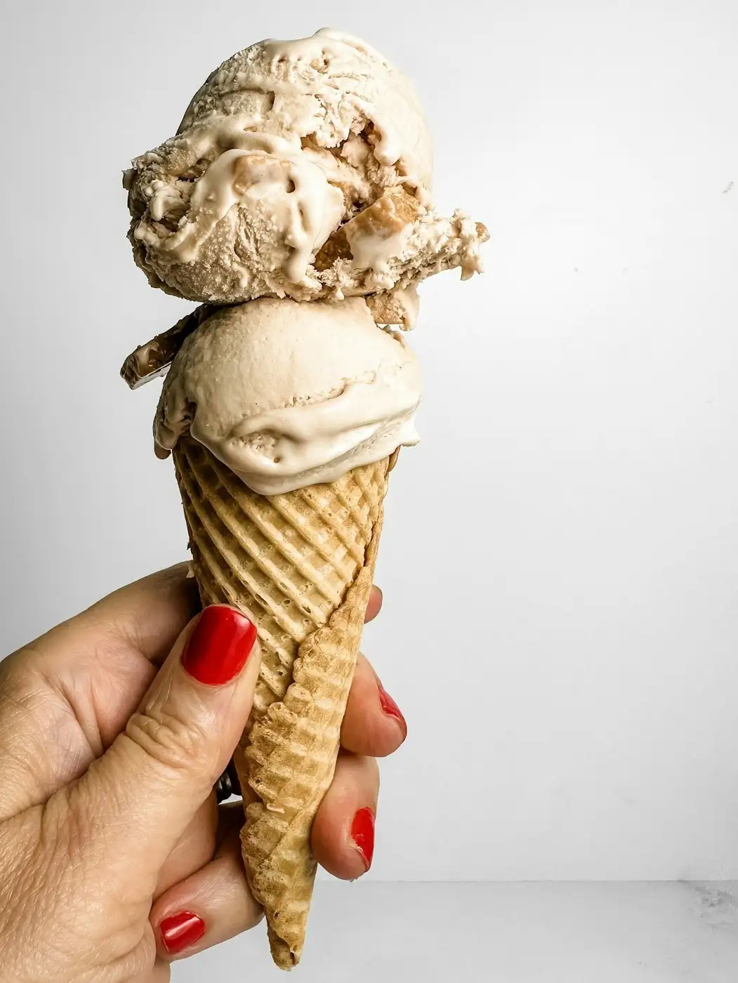 A hand with red nails holding a waffle cone with two scoops of caramel-colored ice cream studded with cake pieces