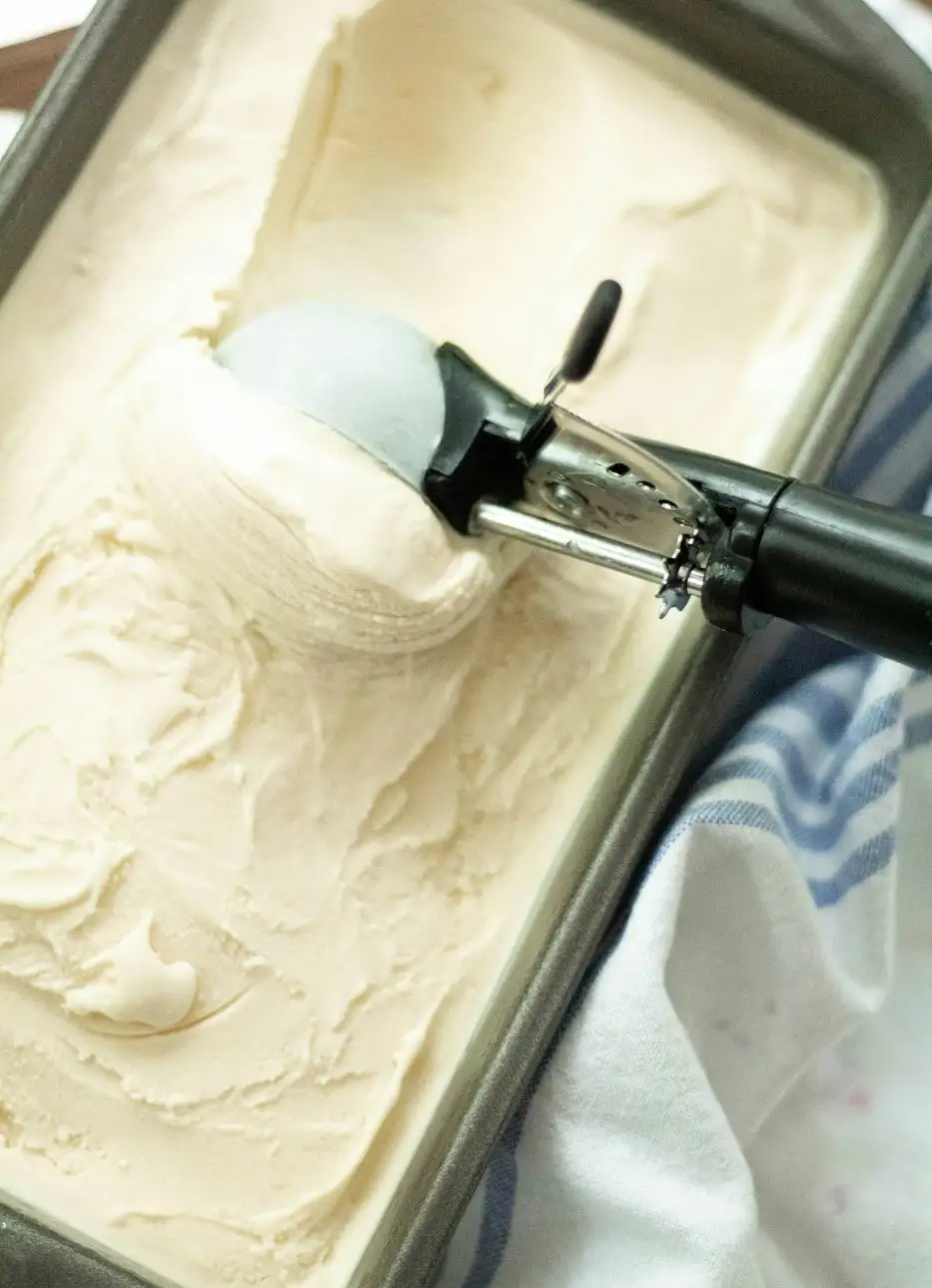A scoop pulling a curl of vanilla ice cream from a full pan