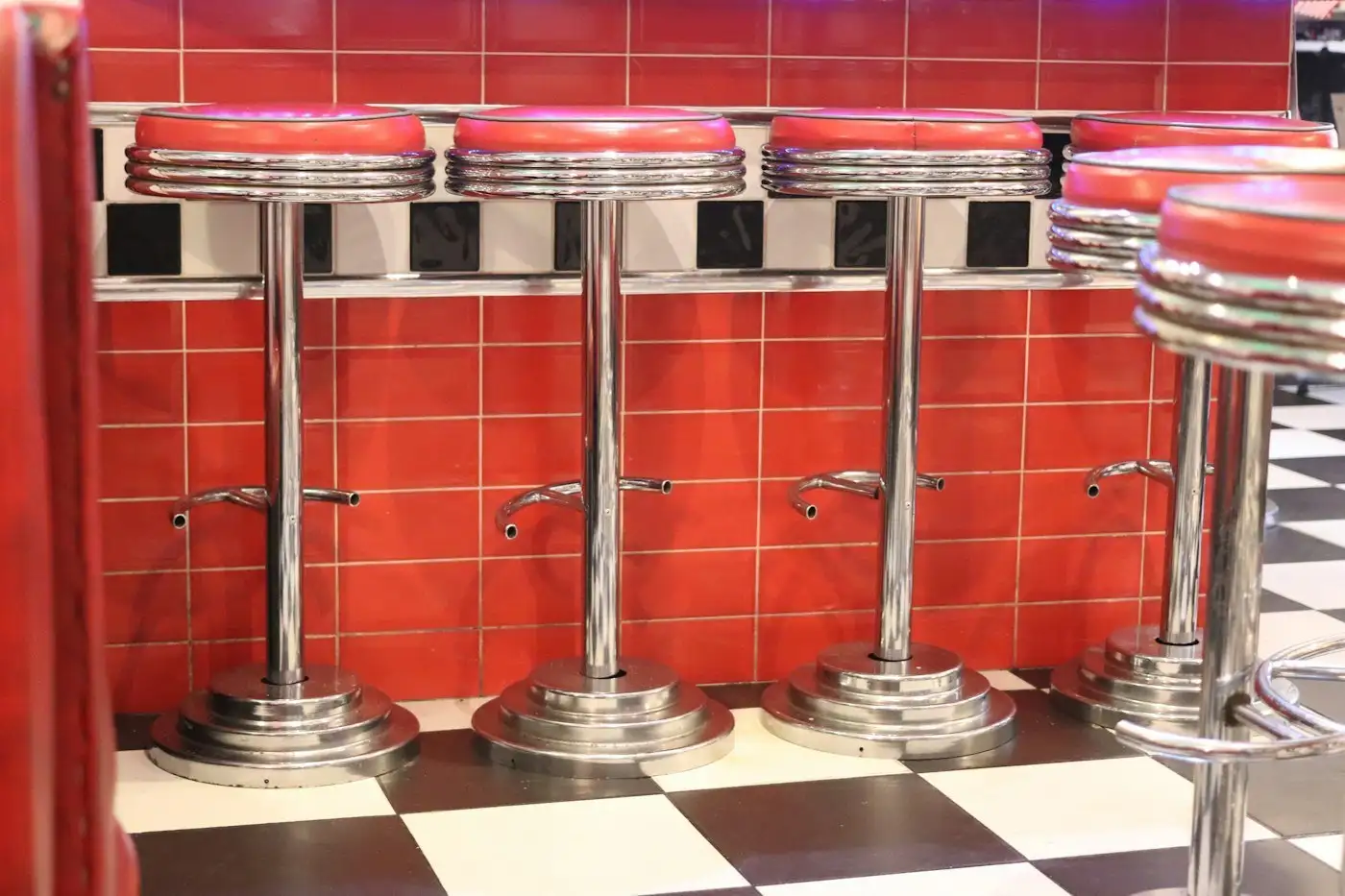 Chrome soda counter stools with red seats against a red tiled wall over a checkerboard floor